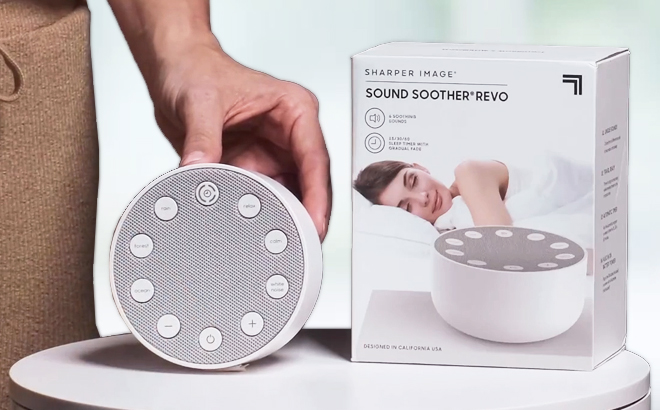 Hand Holding Sharper Image Sound Soother REVO White Noise Machine on a Desk with Box on the Right