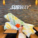 Person Holding 2 Subway Sandwiches inside of a Subway Restaurant