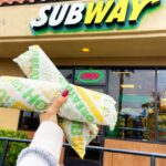 a Hand Holding Two Footlong Subs in Front of a Subway Chain