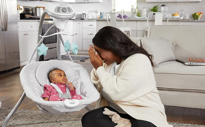 A mother playing with her baby who is resting on a Graco Simple Sway Swing