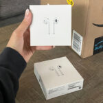 Hand Holding Apple AirPods with Amazon Box in the Background