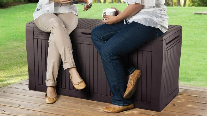 Two Women Sitting on Keter 71 Gallon Resin Deck Box in Espresso Brown