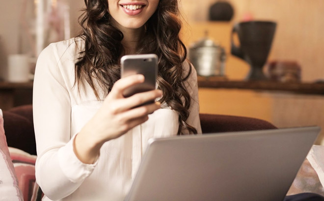 A Smiling Person Sitting in Front of a Laptop and Looking at a Mobile Phone