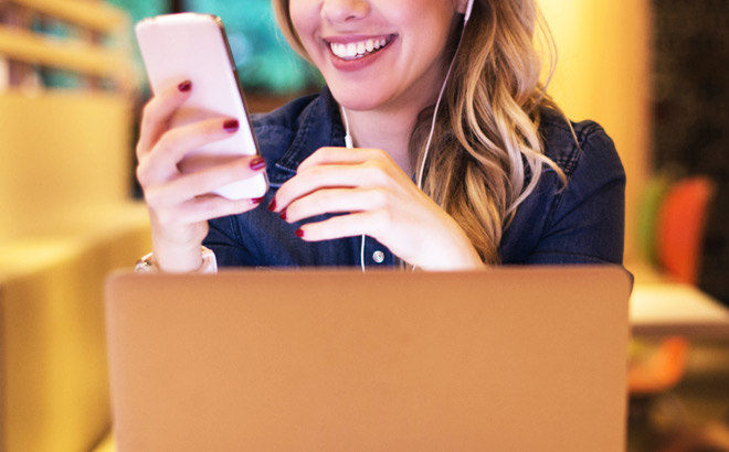 A Smiling Person In Front of a Laptop Looking at Mobile Phone