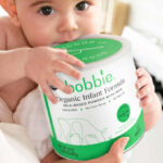 A Baby in Mothers Arms Holding a Can of Bobbie Organic Infant Formula