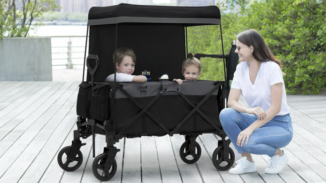 Two kids riding a Little Folks by Delta Children City Wagon Cruiser Stroller with a lady beside them