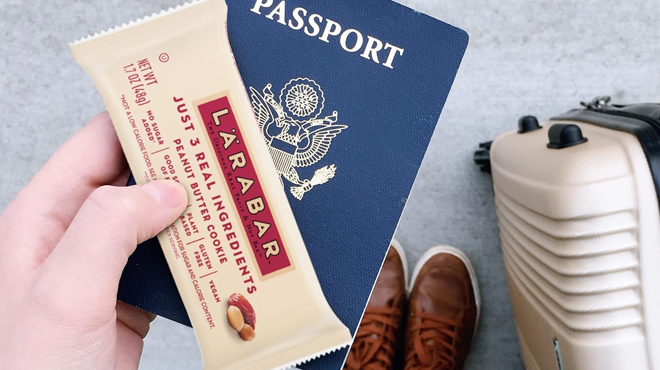 A person holding a Larabar Peanut Butter Cookie and a passport