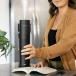 A Woman Putting an Aroma360 Mini Pro Scent Diffuser on a Table next to a Book