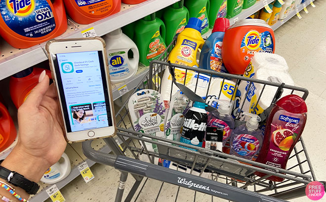 A Hand Holding a Mobile Phone with the Checkout51 App on the Screen and Household Personal Care and Laundry Products on a cart in a Store Aisle